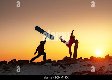 Two happy snowboarders couple man and woman are having fun with snowboards at sunset mountain. Funny silhouettes of young couple are standing in comical poses against sunset yellow and orange sky Stock Photo