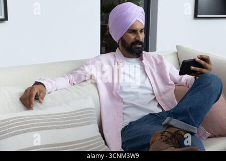 Indian man sitting on sofa in living room holding smartphone, gazing at screen, copy space Stock Photo