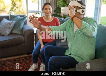 Asian couple sitting on sofa in living room using white vr headset reaching and watching Stock Photo