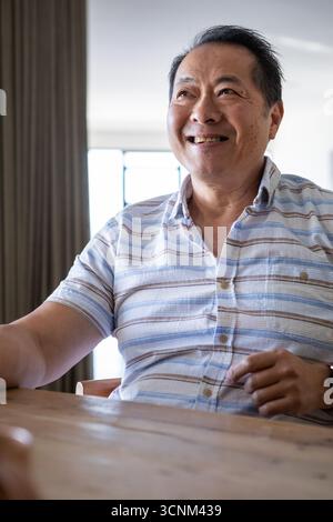 Senior asian man wearing shirt smiling while sitting at home dining table by window light Stock Photo