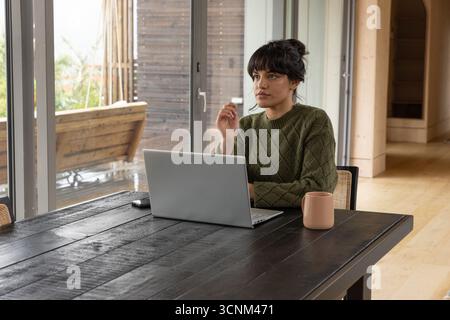 Woman sitting at home office table using laptop, holding smartphone and mug near glass door Stock Photo