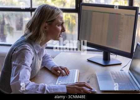 Female office worker typing spreadsheet data at desktop monitor on wooden desk Stock Photo