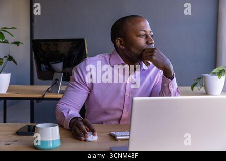 African american man sitting at desk in modern office gazing thoughtfully at laptop with mug Stock Photo