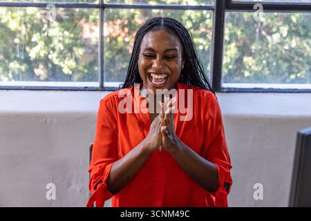 African american woman standing in office by window laughing clasping hands near desk Stock Photo