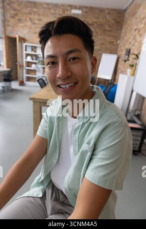 Asian man sitting at desk in loft office smiling while writing on whiteboard Stock Photo