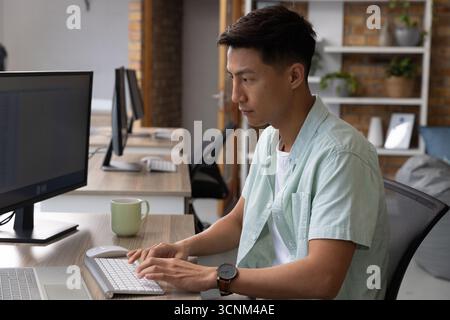 Asian man typing on wireless keyboard at open-plan office desk with laptop, mug, copy space Stock Photo