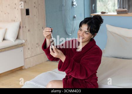 Asian woman sitting in bathrobe examining brown dropper bottle on bed in bedroom, copy space Stock Photo