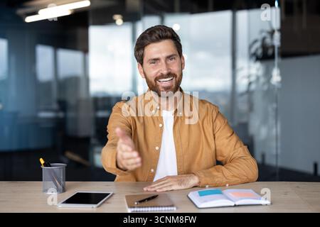 Young businessman looking at camera and extending hand for a friendly handshake, sitting at desk in modern office, concept of job interview, greeting, welcome, or partnership Stock Photo