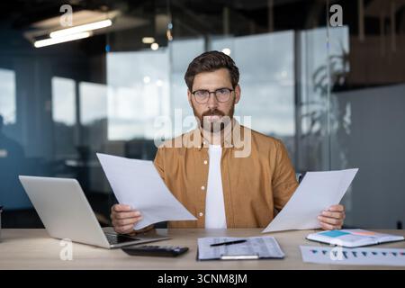 Confident businessman working at a desk in a modern office, holding important documents, analyzing data with a laptop and calculator, demonstrating focus and expertise Stock Photo