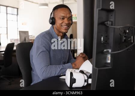 African american man sitting in office wearing shirt, headphones, using computer, copy space Stock Photo