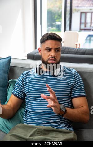 Bearded man gesturing while talking on couch in living room wearing headphones and wristwatch Stock Photo