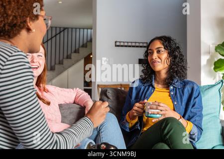 Diverse female friends sitting on couch in living room holding ceramic mug and throw pillows Stock Photo