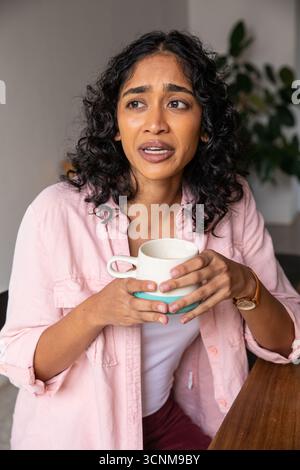 Asian woman sitting at home table wearing wristwatch, holding mug looking worried near plant Stock Photo