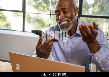 Senior african american man sitting at wooden desk in office holding smartphone, using laptop Stock Photo