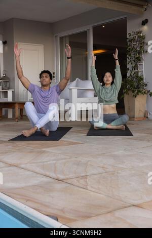 Diverse couple practicing yoga sitting cross-legged raising arms overhead at poolside on mats Stock Photo