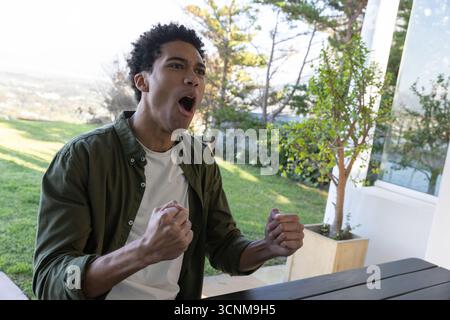 African american man cheering with clenched fists at picnic table on patio overlooking yard Stock Photo