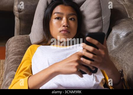Asian teenage female lying on brown leather sofa using smartphone on gray cushion in living room Stock Photo