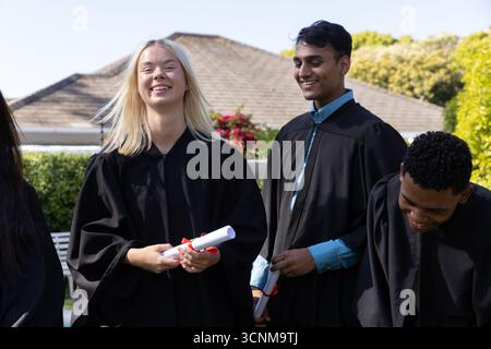 Diverse classmates standing on campus lawn wearing black robes with red ribbon diplomas Stock Photo