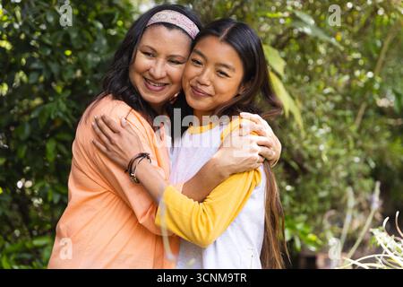 Hispanic mother and Asian teenage daughter hugging in backyard garden with headband and bracelets Stock Photo