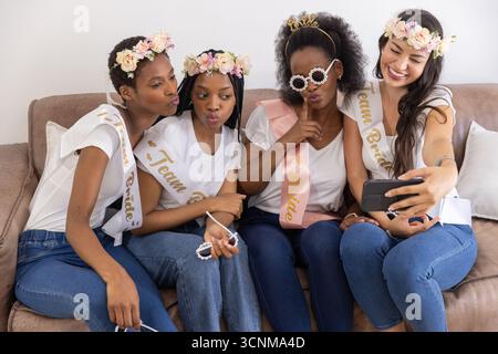 Diverse female friends wearing floral headbands and sashes taking selfie with smartphone on sofa Stock Photo