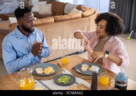 Diverse couple seasoning eggs and giving thanks at home dining table with avocado, orange juice Stock Photo