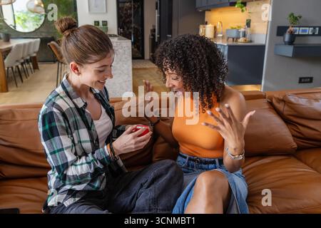 Two women sitting on leather couch offering red ring box in modern living room Stock Photo