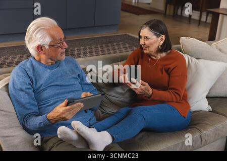 Senior couple sitting on sofa talking while using tablet and smartphone in living room Stock Photo