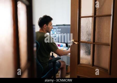 Remote view of focused programmer male working from home office using multiple monitors setup and typing on mechanical keyboard looking to screen Stock Photo