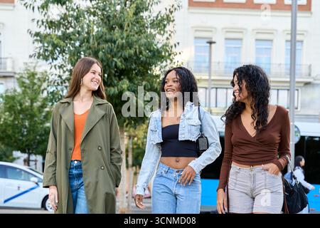 Three diverse young women enjoy a leisurely stroll down a city street, engaging in lively conversation and sharing laughter Stock Photo