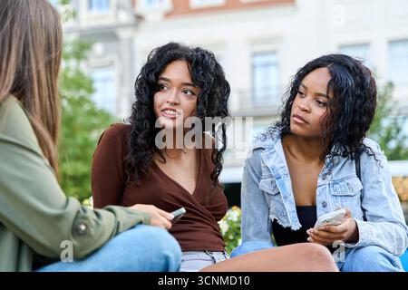 Three young women sitting outdoors in a park, engaging in conversation while browsing their smartphones and enjoying each other's company Stock Photo