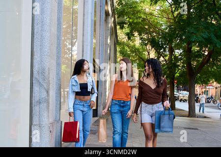 Three diverse young women carrying shopping bags are walking down a city sidewalk, enjoying conversation after a successful shopping spree Stock Photo
