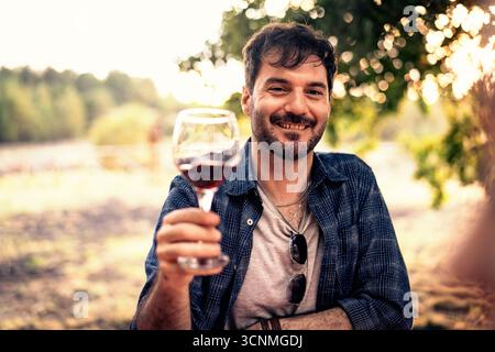 A handsome smiling man makes a toast directly to the camera with a glass of red wine while sitting outdoors in a vineyard during a beautiful golden ho Stock Photo