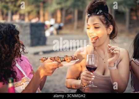 Two happy young female friends are smiling and sharing grilled barbecue skewers from a plate at an outdoor summer picnic with red wine. Stock Photo