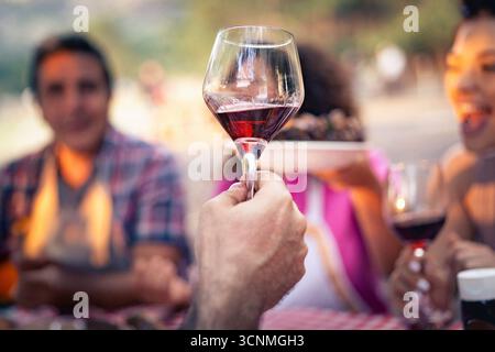 A selective focus shot on a man's hand holding a glass of red wine, with a happy woman laughing in the beautifully blurred background during an outdoo Stock Photo