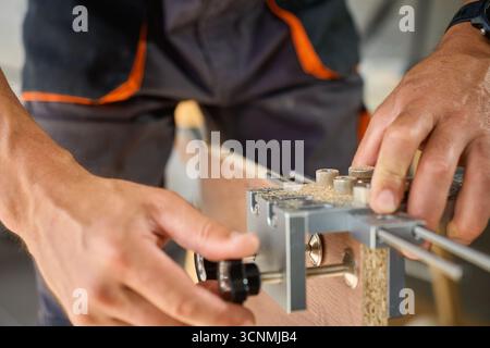 Close-up of carpenter using drilling jig tool for drilling holes in wooden panel. Man assembling furniture in workshop. Concept of woodworking, furnit Stock Photo