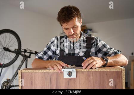 Close-up of carpenter using drilling jig tool for drilling holes in wooden panel. Man assembling furniture in workshop. Concept of woodworking, furnit Stock Photo