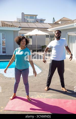 Diverse couple wearing sportswear practicing stretching exercises on yoga mats, backyard pool Stock Photo