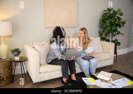 Diverse female colleagues discussing project using laptop, tablet, charts on sofa in living room Stock Photo