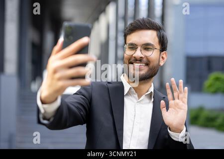Businessman in suit making a video call on his smartphone, smiling and waving to the camera, using mobile technology for communication and connection Stock Photo