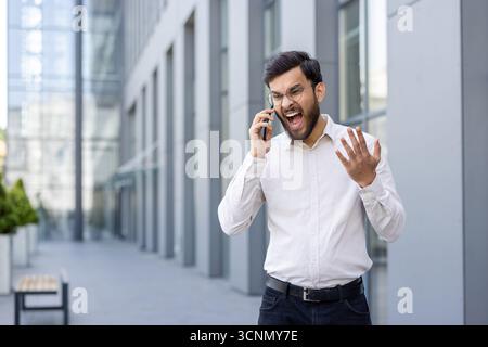 Young businessman yelling into his mobile phone outside an office building, furious and stressed, gesturing angrily while dealing with work pressure, Stock Photo