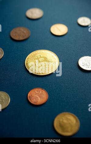 Golden Bitcoin coin among traditional currency coins on blue background highlighting contrast between digital and physical money Stock Photo