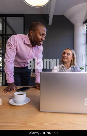 Diverse coworkers discussing project details, leaning on desk with silver laptop in office Stock Photo