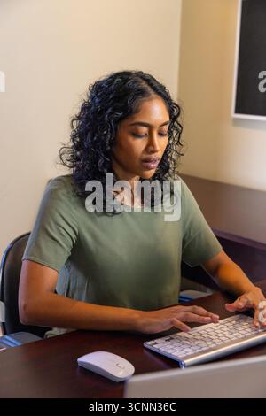 Indian woman typing on keyboard at office desk with wireless mouse focusing on monitor Stock Photo