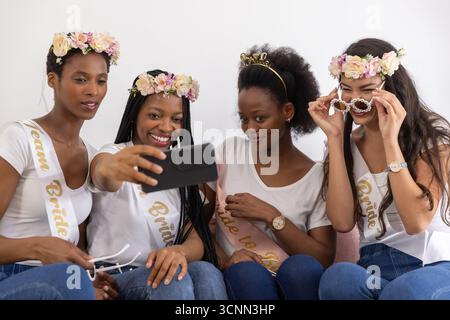 Diverse female friends wearing white tops taking selfie on living room sofa with smartphone, sashes Stock Photo