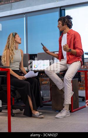 Diverse coworkers reviewing printed charts by red metal desk at office using laptop and smartphone Stock Photo