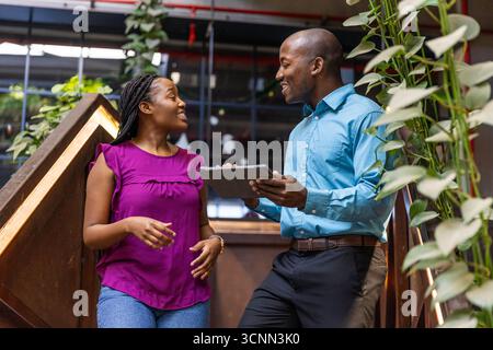 African american colleagues standing on stair with plants discussing data on tablet Stock Photo