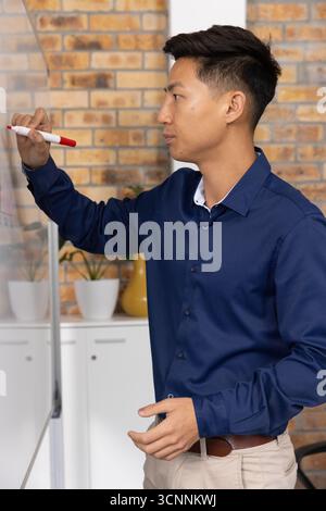 Asian man writing on whiteboard with red-tipped marker in office meeting room with potted plants Stock Photo