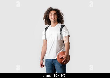 Young man with curly hair holding basketball and wearing backpack isolated on white background. High quality photo Stock Photo