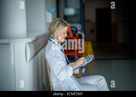 Young female doctor with short hair in sitting on bench in hospital corridor, reviewing documentation. Stock Photo