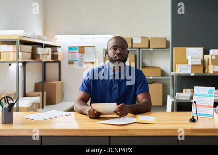 Portrait of young adult Black man sitting at desk holding documents, looking into camera, surrounded by packages and paperwork in warehouse or shipping office environment Stock Photo